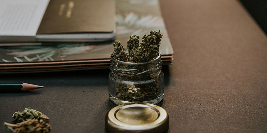 dried green kush cannabis inside a glass container on top of a table beside a pencil and a notebook